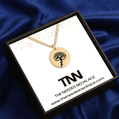 Gold necklace with a flower pendant in a box labeled 'The Needed Necklace' on a blue fabric background.